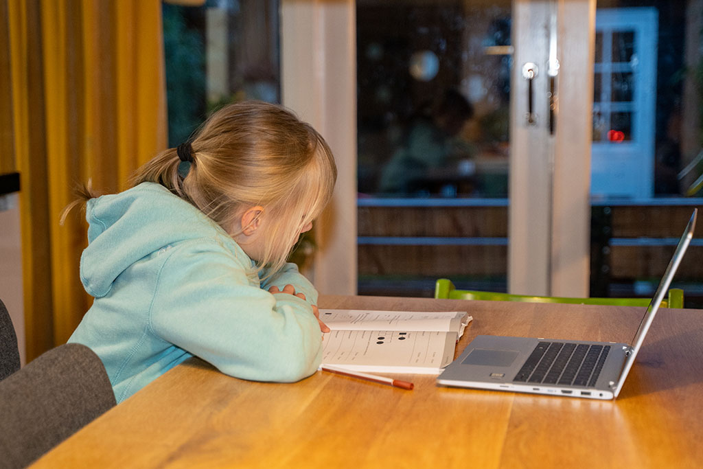 Een leerling werkt thuis aan huiswerk aan tafel met de laptop en een schoolboek.